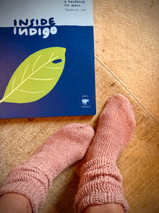 Book title "Inside Indigo' by Julia Tabakhova green leaf on a blue background, beside pink indirubin dyed socks. 